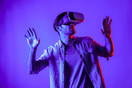 Excited man wearing VR goggle to exploring and enter in virtual program. Skilled gamer touching in metaverse while wearing casual cloth and standing at neon light background. Lifestyle. Deviation.