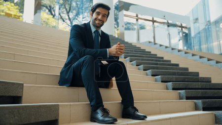 Photo for Smiling skilled businessman looking at camera while sitting on stairs. Young professional project manager smile at camera while holding mobile phone at outdoor with blurred background. Exultant. - Royalty Free Image