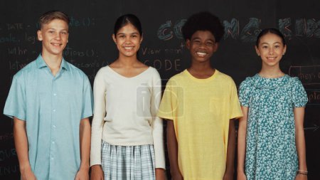 Téléchargez les photos : Groupe d'adolescents multiculturels intelligents regardant la caméra au tableau noir. Heureux enfants diversifiés se tenant dans la classe de technologie STEM avec code d'ingénierie, idée de programme rapide ou de planification. Édification. - en image libre de droit