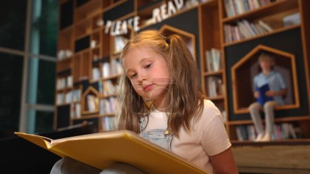 Photo for Young smart caucasian girl picking reading a book while sitting at library. Clever child learning, studying, open a books at library. Attractive kid turning page with blurring background. Erudition. - Royalty Free Image