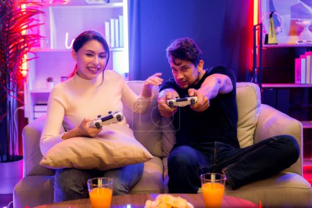 Photo for Couple joyful funny players playing video game on TV using joysticks, fighting winner trying to disturb when focusing level challenge in red neon light studio at comfy living home place. Postulate. - Royalty Free Image