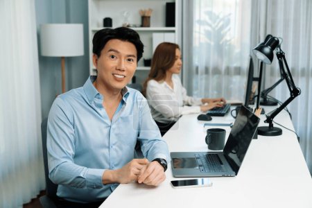 Photo for Profile smart Asian businessman looking at camera while holding coffee cup to pose at modern office. Blurry background woman colleague working on pc to check email marketing report or reply. Infobahn. - Royalty Free Image