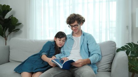 Happy daughter lean on father shoulder while listening story telling. Young asian schoolgirl looking ar dad while reading story book together and sitting at sofa. Family recreation concept. Pedagogy.