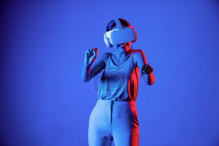 Foto de Soporte femenino inteligente rodeado de luz de neón que usa auriculares VR que conectan tecnología de comunidad ciberespacial metaversa y futura. Mujer elegante disfrutar de jugar juegos de carreras de coches en el mundo meta. Alucinaciones. - Imagen libre de derechos