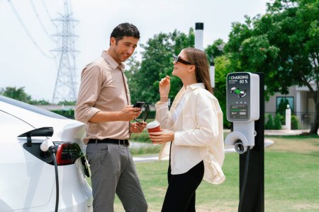 Junge Paare mit Kaffee bezahlen für Strom, während sie an einer an das Stromnetz angeschlossenen Ladestation die Elektroautobatterie aufladen. Sinnvoll
