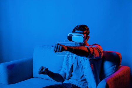 Photo for Happy man drive a car movement while wear VR glasses and sitting at sofa with neon light background. Smart gamer playing sport game while moving driving car gesture by using smart goggle. Deviation. - Royalty Free Image