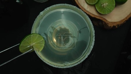 Téléchargez les photos : Macrographie, une vue du haut vers le bas d'une margarita rafraîchissante est présentée dans un verre orné d'une tranche de citron vert, le tout sur fond noir. Chaque plan rapproché du cocktail et du citron vert. Comestible. - en image libre de droit