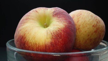 Foto de Close-up muestra una manzana fresca de color rojo brillante y crujiente enclavada cómodamente en un tazón de vidrio. El fondo negro crea un escenario dramático, destacando la piel impecable y lisa de las manzanas. Comestible. - Imagen libre de derechos