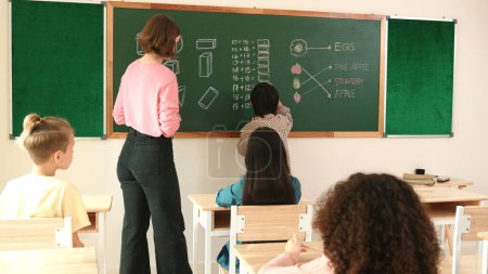 Photo for Smart teacher checking cute student homework while explaining idea at class. Attractive instructor walking around classroom while looking at notebook and talking about Math theory to child. Pedagogy. - Royalty Free Image