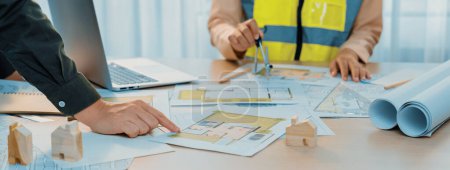 Photo for Skilled engineer uses divider to measure the blueprint while architect pointing the mistake point in the building blueprint on table with architectural document scatter around. Close up. Delineation. - Royalty Free Image