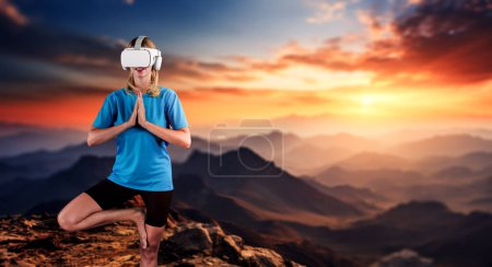 Foto de Chica caucásica practicando yoga mientras usa camisa con la montaña puesta del sol. Mujer inteligente feliz haciendo yoga posando mientras está de pie en el acantilado y usando gafas VR para experimentar la sensación de calma. Artilugio. - Imagen libre de derechos