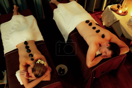 Téléchargez les photos : Massage aux pierres chaudes au salon de spa dans un complexe de luxe avec lumière chaude aux bougies, un couple heureux profitant d'un massage aux pierres de basalte spa glisse sur le corps avec une chaleur apaisante. Tranquillité - en image libre de droit