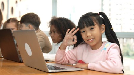 Téléchargez les photos : Smart fille asiatique travaillant sur ordinateur portable faire ok ou ok et regarder la caméra. Divers enfants heureux codant ou programmant le code ou l'invite d'ingénierie en utilisant l'ordinateur dans la classe de technologie STEM. Erudition. - en image libre de droit