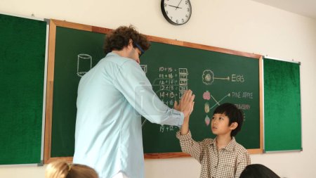 Photo for Attractive boy writing answer while teacher give high five to encourage child. Attractive student walking back to his seat while teacher applause and clap hand to schoolboy at Math lesson. Pedagogy. - Royalty Free Image