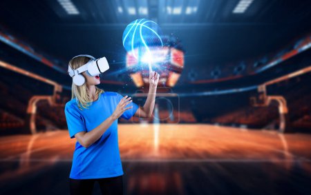 Téléchargez les photos : Fille pointant ou filant basket tout en utilisant des lunettes VR et visiter le stade de sport. Femme caucasienne regardant la balle tout en portant des lunettes de réalité visuelle et debout à l'arène de basket-ball. Machines et appareils. - en image libre de droit