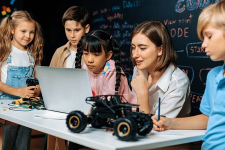Photo for Skilled kids learning to program remote car. Schoolgirl in pink cloth use laptop for coding. Other child watch her code while teacher watch her. motherboard and electric wire also on table. Erudition. - Royalty Free Image