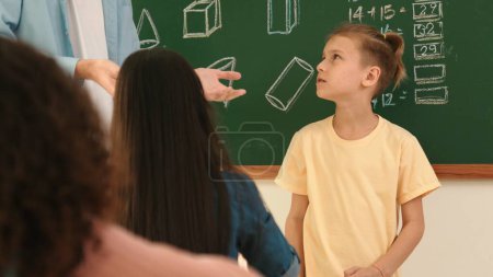 Professionelle Lehrer sprechen und erklären Idee zu asiatischen Kind während des Unterrichts. Kaukasischer Student hört Lehrer zu, während er vor einem Klassenzimmer steht, in dem eine Mathestunde an der Tafel geschrieben wird. Pädagogik.