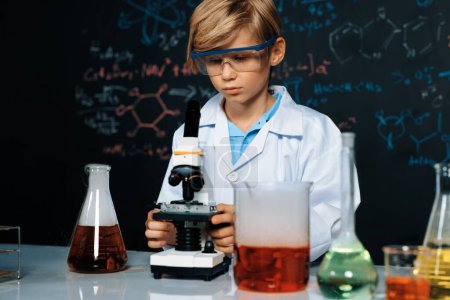 Téléchargez les photos : Un écolier aux cheveux blonds en blouse de laboratoire apprend la science de la technologie chimique en classe STIM. L'étudiant regardant objet au microscope. Sur la table mettre beaucoup de fiole. Erudition. - en image libre de droit
