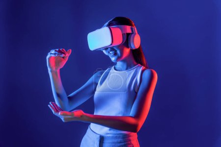 Téléchargez les photos : Smart Female debout entouré de néon lumière usure VR casque connectant métaverse, technologie futuriste de la communauté du cyberespace, en utilisant les deux mains interagissent avec l'objet virtuel généré. Hallucination. - en image libre de droit