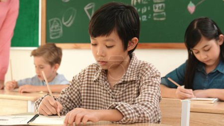 Téléchargez les photos : Asiatique attrayant garçon prendre une note tandis que l'étudiant écrit réponse dans la feuille de réponse. Étudiant multiculturel faisant du travail en classe ou des tests tout en enseignant caucasien vérifiant les devoirs des élèves en classe. Pédagogie. - en image libre de droit