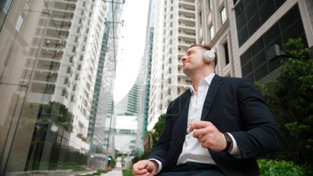 Photo for Low angle camera of project manager listening relaxing music while move to song and clap hands. Happy caucasian businessman enjoy listening music while sitting at bench at green urban city. Urbane. - Royalty Free Image