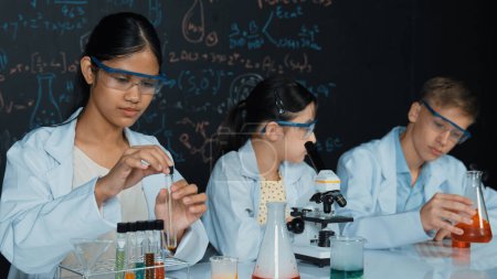 Photo for Cute girl looking under microscope while student doing experiment at blackboard with theory written. Young scientist inspect colored solution at table with experimental equipment placed. Edification. - Royalty Free Image
