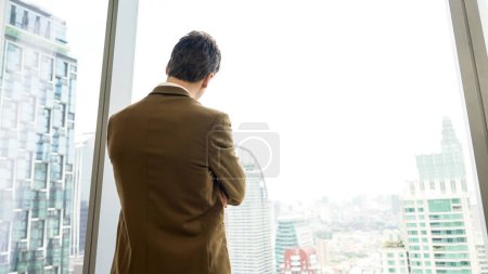 Téléchargez les photos : Arrière vue ambitieux homme d'affaires debout dans le bureau décoré regardant par la fenêtre sur l'horizon du paysage urbain. La détermination et l'ambition d'affaires poussent la carrière d'affaires vers un avenir brillant - en image libre de droit