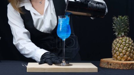 Macrografía, experimente el arte de una mano de camareros expertos mientras vierten expertamente un cóctel Blue Hawaii en vidrio sobre un llamativo fondo negro. Cóctel. Primer plano de tiro. Comestible.