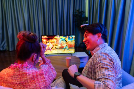 Photo for Couple gamer with joysticks playing fighting video game on tv screen, man getting win challenge level at back with girlfriend, sitting sofa at neon light color living room at modern home. Infobahn. - Royalty Free Image