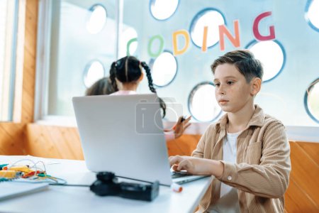 Photo for The schoolboy in classroom sitting and learning about engineering remote control with laptop while other child watch outside. Circuit wire, motherboard and notebook put on desk messily. Erudition. - Royalty Free Image