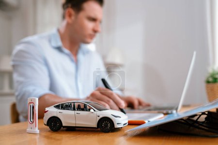 Focus scale EV car and charging station mockup model on blurred background of automotive design engineer working on designing and looking for improvement for EV car design on laptop. Synchronos