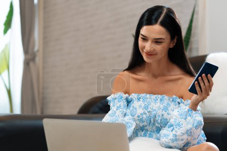 Photo for Young woman using laptop, happily browsing the internet or making online purchases on sofa at home. Modern woman enjoying her domicile lifestyle by studying online or simply relaxing. Blithe - Royalty Free Image