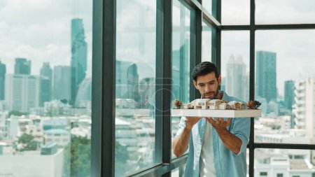 Photo for Businessman in casual outfit holding house model while checking house construction. Architect engineer inspect building model while standing near window with skyscraper. Engineering. Tracery - Royalty Free Image