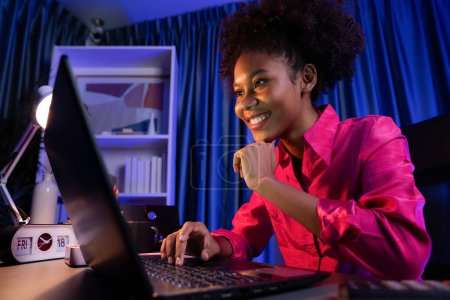 Téléchargez les photos : Blogueuse africaine portant une chemise rose avec un visage heureux, regardant un ordinateur portable à l'écran avec un projet de réussite valorisé ou obtenir une bourse d'études. Concept de travail d'expression gai de la maison. Tastemaker. - en image libre de droit