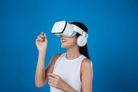 Téléchargez les photos : Femme intelligente debout avec fond bleu portant casque VR connectant métaverse, technologie futuriste de la communauté du cyberespace. Femme élégante excitée de voir généré des paysages virtuels. Hallucination. - en image libre de droit