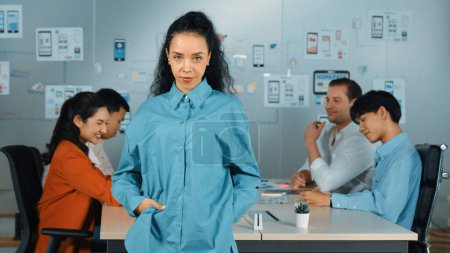 Hispanic professional businesswoman standing while looking at camera in creative business meeting. Beautiful confident leader posing while diverse business people discussion about plan. Manipulator.