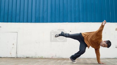 Téléchargez les photos : L'homme hispanique tend les bras et danse la rue en dansant devant le mur. Motion shot du danseur ou chorégraphe élégant en tenue décontractée pratiquant la danse dans le style hip hop. Sport de plein air 2024. Endeavor. - en image libre de droit