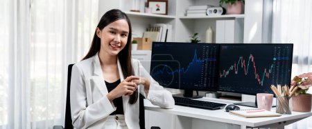 Foto de Perfil de la joven mujer de negocios asiática sonriendo en la cara feliz con traje blanco sentado silla contra el mercado de valores dinámico gráfico de inversión en la PC que muestra la pantalla en el escritorio en la oficina moderna. Estratagema. - Imagen libre de derechos