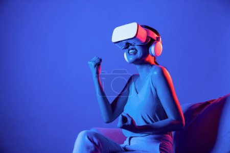 Photo for Smart Female sitting on sofa surrounded by neon light wear VR headset connecting metaverse, futuristic cyberspace community technology, watch movie with cheerful action and happy face. Hallucination. - Royalty Free Image