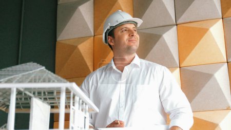 Photo for Portrait of professional project manager holding project plan with house model at table. Professional architect engineer wearing safety helmet while standing posing with confident. Manipulator. - Royalty Free Image