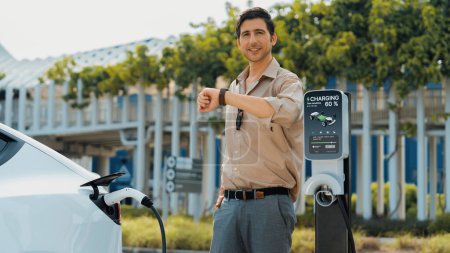 Photo for Young man checking time on smartwatch while EV charger to recharging battery from charging station in parking lot. Rechargeable EV car for sustainable eco friendly urban travel. Panorama Expedient - Royalty Free Image