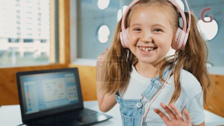 Smiling girl looking while waving hand at camera with laptop placed on table. Child wearing headphone smiling while laptop screen show system programing or coding program in STEM class. Erudition.