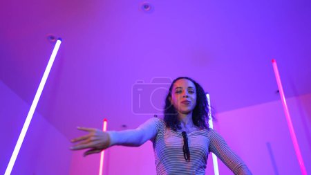 Photo for Low angle camera of hispanic dancer looking at camera and dancing to city pop music. Professional attractive hipster moving to hip hop song with purple led or neon light. Stylish cloth. Regalement. - Royalty Free Image