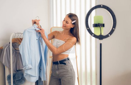 Photo for Young woman making fashion video content for social media. Blogger smiles to camera using selfie stick and light ring while making persuasive online clothing sell to audience or followers. Unveiling - Royalty Free Image
