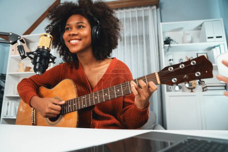 Téléchargez les photos : Chaîne hôte en musicien de jeune afro-américain jouant de la guitare avec le chant, diffusion sur ordinateur portable en studio. Décoration de l'équipement des casques et microphone enregistreur. Tastemaker. - en image libre de droit
