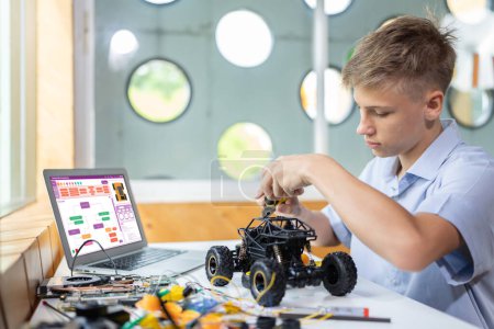 Photo for Blonde hair boy carefully using pliers fixing robotics car while learning technology in STEM class. On table put laptop, controller, electric wire, battery charger, and robotic vehicle. Edification. - Royalty Free Image