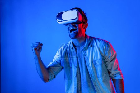 Téléchargez les photos : Heureux homme excité de regarder à la compétition sportive au monde virtuel tout en portant un casque VR à fond clair néon. Adolescent beau entrer dans la technologie de l'innovation future en utilisant des lunettes. Déviation. - en image libre de droit