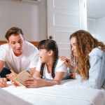 Bedtime story with childhood storytelling, mom and dad reading a fantasy book together to their little young girl in cozy and comfortable bedroom. Modern family happy time. Synchronos