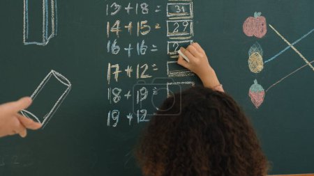 Rückansicht eines afrikanischen Schülers beantwortet die Frage, während er an Bord schreibt. Lehrer schauen Grundschülern beim Mathe-Quiz vor der Klasse zu, während intelligente Kinder im Mathe-Unterricht dabei sind. Pädagogik.