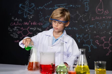 Photo for Blonde hair schoolboy in laboratory wear lab coat stand and learn science of chemistry technology in STEM class. The student pour liquid down to glass beaker. On the table put many flask. Erudition. - Royalty Free Image
